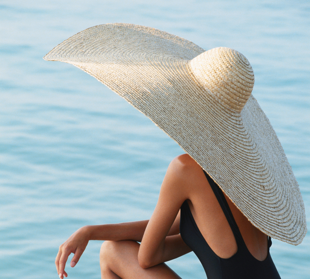 Woman sitting alongside sea water in a black bathing suit and large over sized sun hat