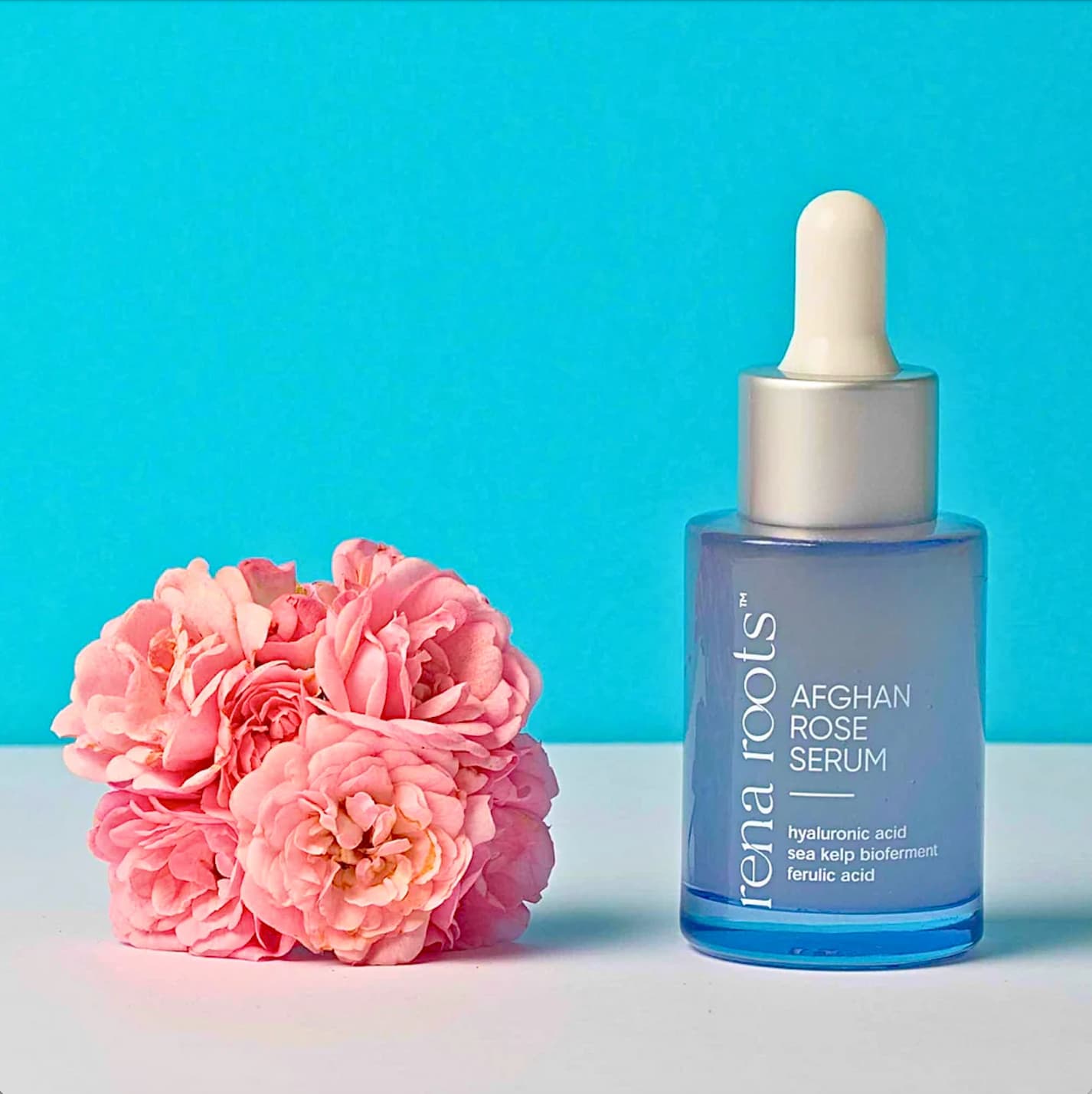 A bottle of  Rena Roots Afghan Rose Serum with dropper and pink roses on a blue background