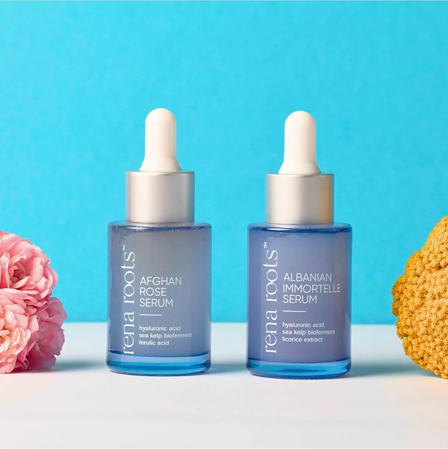 Two blue Rena Roots Serum bottles with droppers on a light blue background, surrounded by flowers.