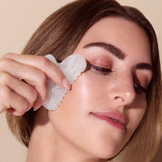 Woman using a Rena Roots White Jade Gua Sha tool on her face against a beige background