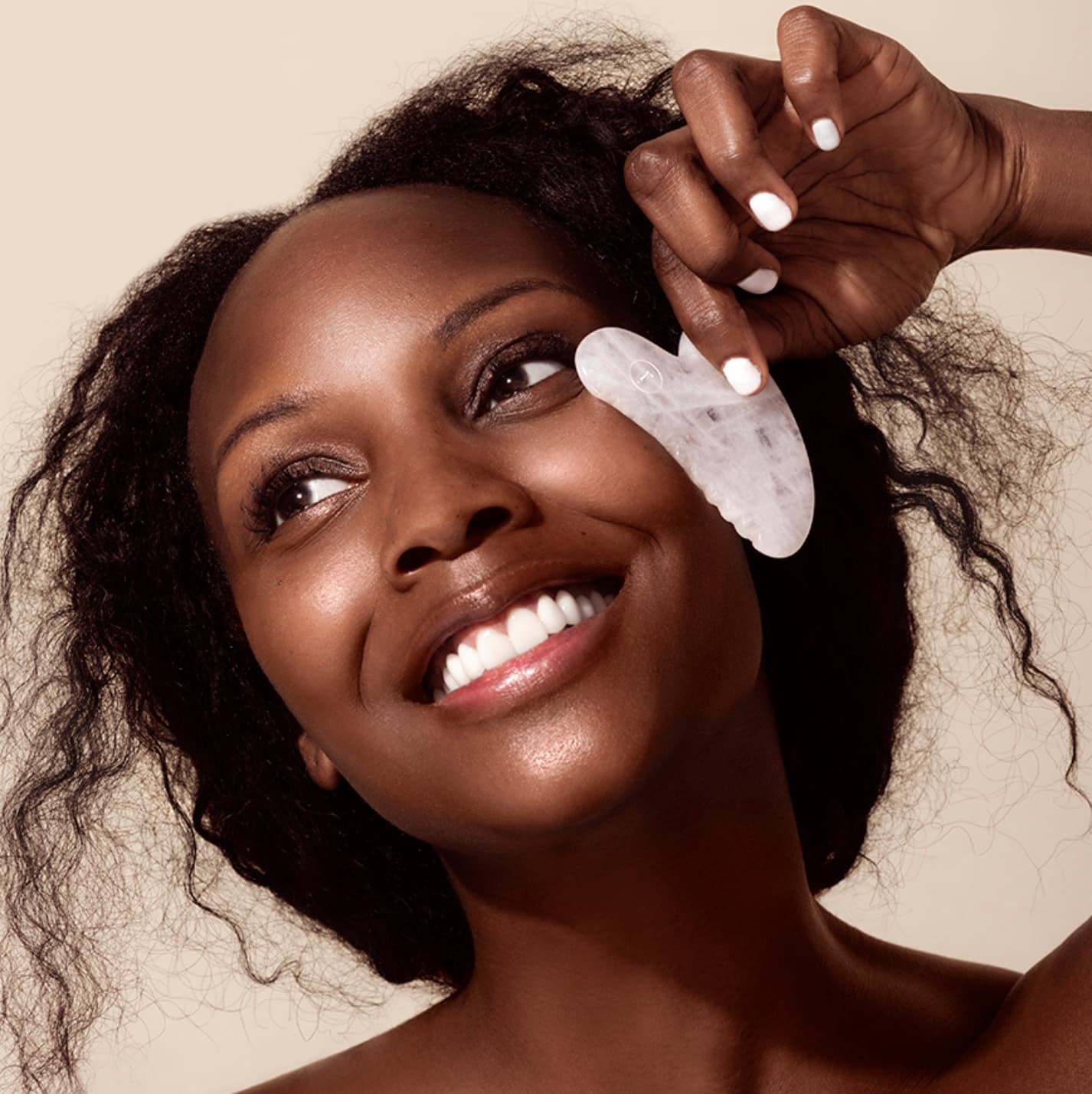 Woman using a Rena Roots White Jade Gua Sha tool on her face against a beige background