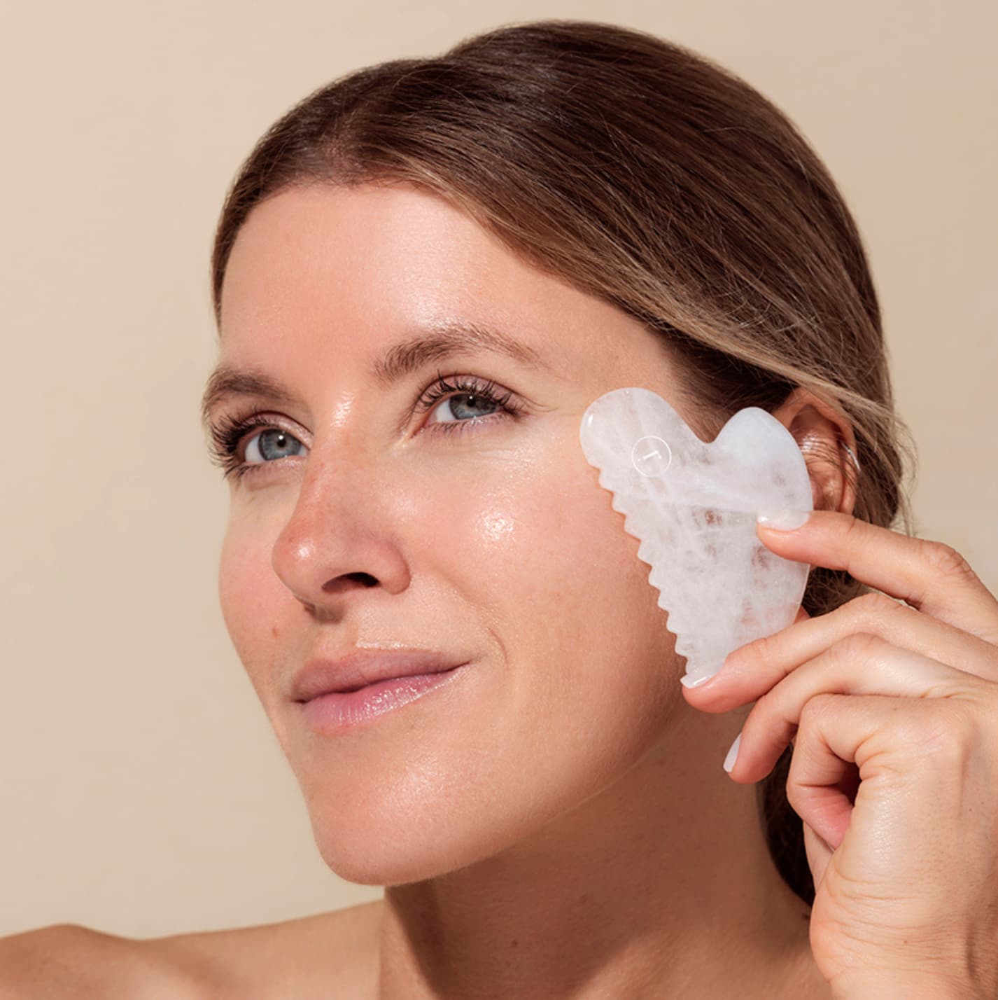 Woman using a Rena Roots White Jade Gua Sha tool on her face against a beige background